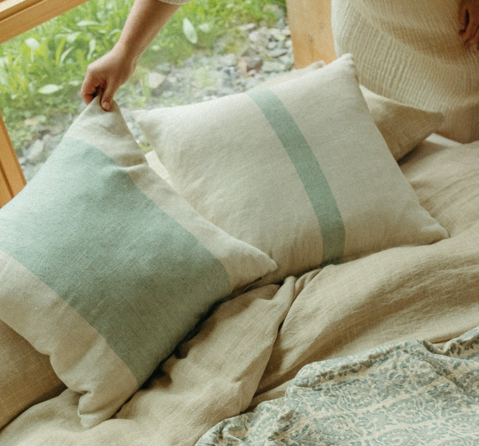 A beige linen pillow with a green stripe, displayed on a bed with a matching linen bedsheet.