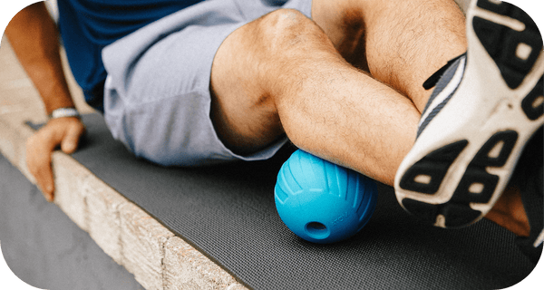 Person using a blue massage roller under their calf while seated outdoors on a mat, demonstrating recovery and wellness with the RAD ATOM | RAD product.