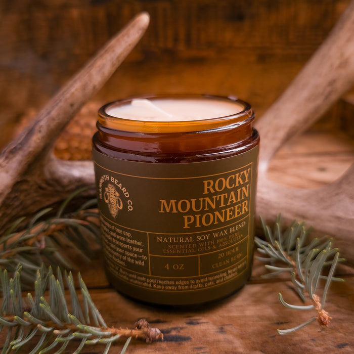 4 oz Rocky Mountain Pioneer soy wax candle with lid off, revealing creamy wax, displayed with antlers and pine greenery on a wooden surface.