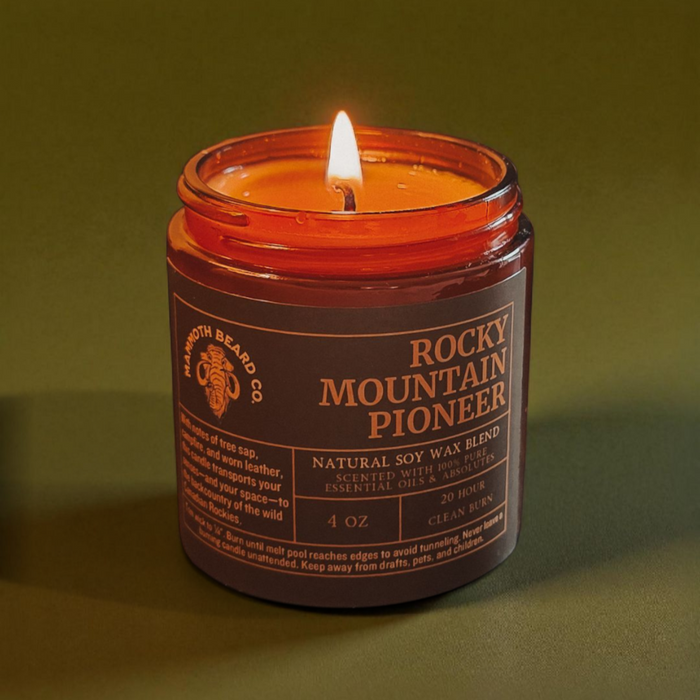 Rocky Mountain Pioneer natural soy wax candle lit, amber glass jar on an olive-green background, showcasing clean burn and warm flame.